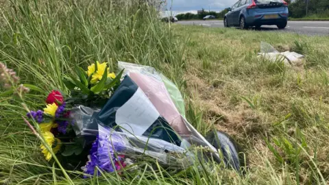 Flowers by the side of Newark Road, North Rauceby, where two teenager suffered fatal injuries in a car crash. There are multiple bunches wrapped in cellophone placed in long grass. In the top right corner of the photo you can see a car driving along the road.