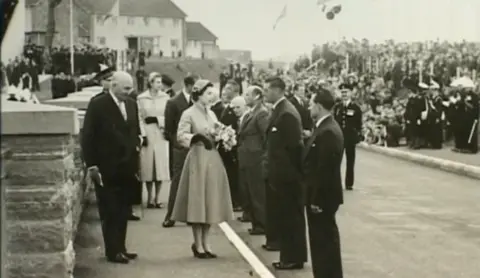 Bristol Water Black and white shot of the late Queen Elizabeth II talking to dignitaries in suits. There are crowds of people in the background.