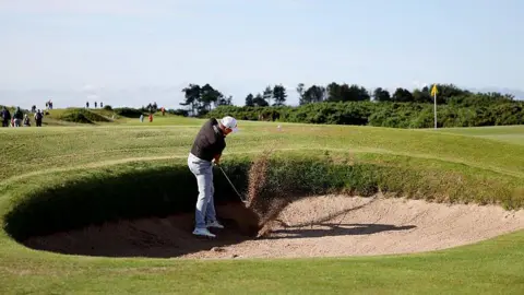 A wide shot of a golfer wearing a dark polo shirt, light grey trousers and a white cap chipping a golf ball out of a bunker. Sand can be seen spraying up where his club and the ball made contact and the ball has lifted just out of the bunker. In the distance the yellow flag marking the hole can be seen as well as some scrubland and, further up the image, the blue sky.