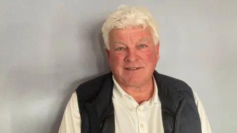 A man with white slightly curled hair is in a white shirt and blue gilet and stands in front of a plain grey wall.