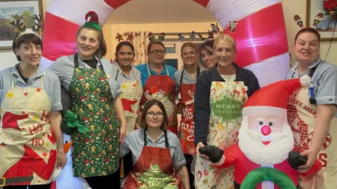 A group of nine women and a large inflatable candy cane and Santa. The women are wearing festive aprons with the words "Merry Christmas" and "Ho Ho Ho" on them.