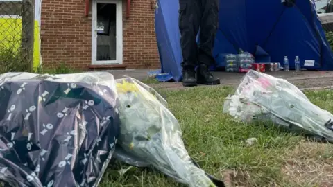 Sam Read/BBC Three bouquets of flowers, on the ground, by grass, with police feet in the distance. The flowers are covered in plastic.
