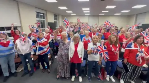 A choir stands in a group looking at the camera and waving union jack flags and many wearing the same blue white and red colours.