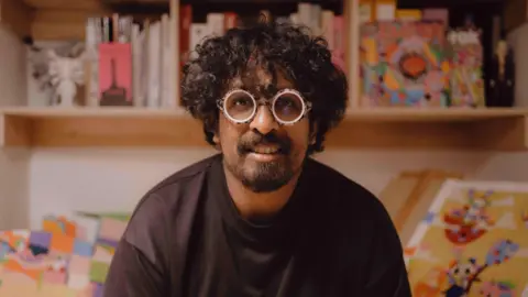 Portrait image of Murugiah looking straight into the camera with a neutral expression. He is wearing a black T-shirt, glasses, has curly black hair and a short beard. His background is made up of out-of-focus books and illustration pieces he has made.