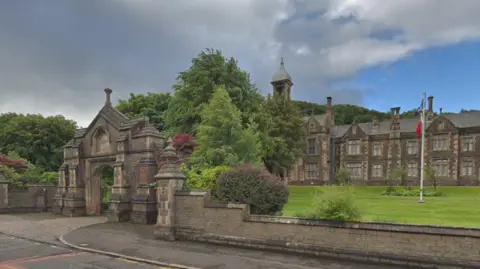A large care home building, with a gated entrance and several large trees surrounding it