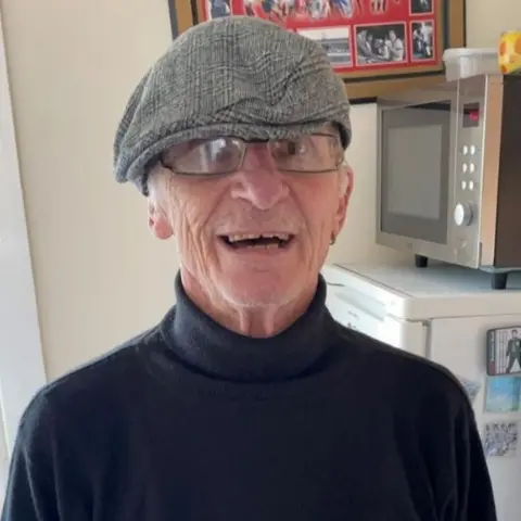 Family handout A man, with short grey hair protruding from from under a flat cap, smiles at the camera. He is standing in a kitchen with a microwave oven and a fridge behind him.