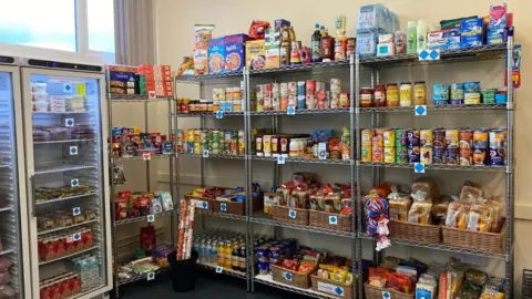 There are shelves full of tinned items, baskets of bread and boxes of cereal inside the pantry. There are fridges with meat products to the left of the pantry. 