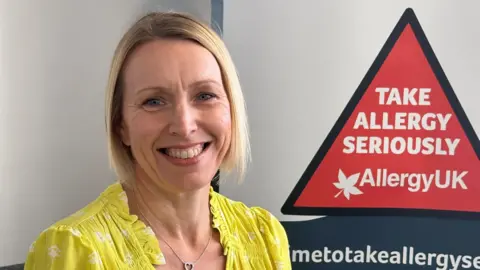 Allergy UK Simone Miles sitting looking direclty at the camera and smiling. She is wearing a yellow blouse. Behind her is a sign that reads "Take Allergy Seriously". 
