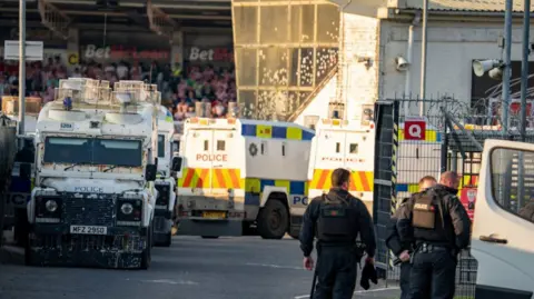 Aodhan Roberts Three police officers are standing next to a vehicle with a door open. A crowded stadium is behind them. Several police land rovers are parked. 
