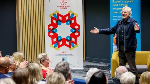 Larry Lamb wearing a black button up shirt with white buttons and a dark navy jacket. He has black trousers on. He has his hands spread out in front of him and he is speaking to a large crowd who are seated and he is standing. Behind him a yellow and grey arm chair 