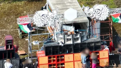 Avon and Somerset Police A close up of one of the makeshift stages with lots of speakers coming out of the back of a large white van. There are metal gantries and scaffolding with two large wasp artworks on either top corner.