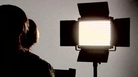 A woman sitting in front of a bright light for a TV interview.