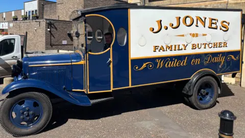 Luke Deal/BBC A pre-war blue van, with yellow livery around its doors, windows and edges. A sign painted on the side of the rear of the van says "J.JONES, FAMILY BUTCHER, Waiting on Daily".
