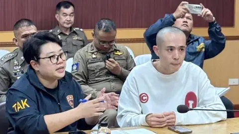 EPA-EFE Wang Xing with a shaved head sitting at a table. Beside and around him are Thai officials in uniform.