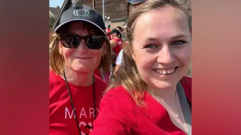 Jen Craft Two women both wearing red; the woman on the left is wearing sunglasses and a sun hat. She has short brown hair and smiling at the camera. Another woman has blonde hair and is also wearing a red top and a small silver necklace.
They are both smiling at the camera 