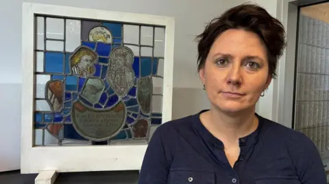 BBC/Cathy Killick A woman with short dark hair, brown eyes and wearing a blue collarless shirt stands next to a panel of stained glass. It shows detailed faces and portions of blue and orange glass.