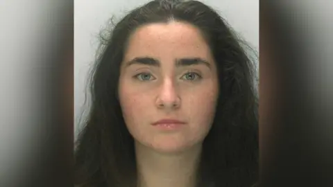 A police custody mugshot of Liberty Mitchell. She has long, dark hair and is staring at the camera with a blank expression