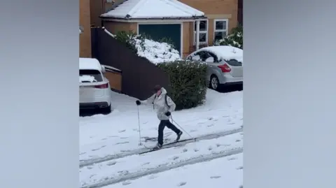 A man skiing down a residential street. He is wearing a grey jacket and black trousers.