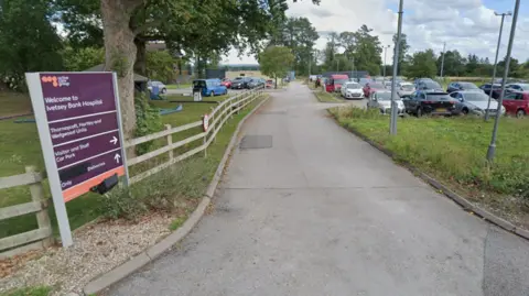 The entrance to a hospital car park with a wooden fence along a grassed area on one side, next to which a purple direction sign says "Welcome to Ivetsey Bank Hospital" and directs visitors to various units, parking and deliveries.