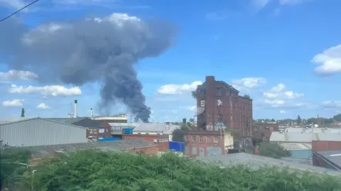 Black smoke billows into a bright blue sky, with industrial buildings