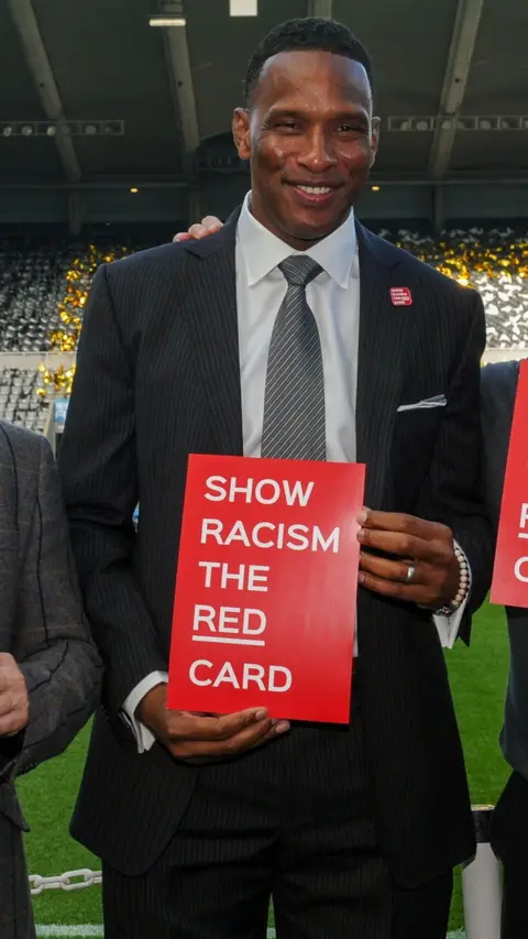 Shaka Hislop stands in front of St James' Park's East Stand in a dark pinstripe suit, holding a red sign that reads Show Racism the Red Card. 
