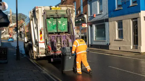 Getty A bin attendant wheels two black bins up to the back of his lorry. He wears bright orange protective clothing with LBC on the back. It is on a high street with traditional buildings on either side. 