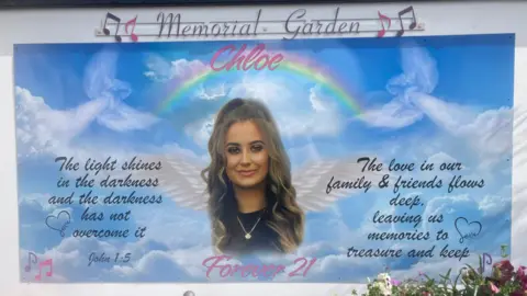 A memorial garden for Chloe. Her face is in the centre, she has long brown curly hair and is wearing foundation, mascara and lipstick. She is wearing a fine necklace and is smiling with her mouth closed. She has white wings coming from her just below her face. There is a rainbow above her with her name 'Chloe' above her face and then 'Forever 21' below her face. On the left hand side of her face, in black writing reads John 1:5 'The light shines in the darkness and the darkness has not overcome it.' On the right hand side of her face, in black writing reads 'The love in our family & friends flows deep, leaving us memories to treasure and keep'. The background is a blue sky with clouds and angels subtly drawn and black hearts and pink music notes. 
