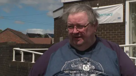 Christopher Morris has light coloured hair and is wearing glasses and a blue and purple t-shirt with a white logo on it. He is speaking outside the medical centre.