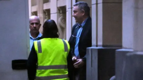 A middle-aged man in handcuffs is led out of a building by a woman in a fluorescent jacket
