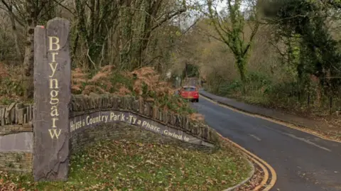 A tree-lined country road curves gently through a wooded landscape at the entrance to Bryngarw House and Country Park. In the foreground, a tall vertical stone marker carved with the word “Bryngarw” stands beside a curved stone wall bearing bilingual Welsh and English signage for the park. Brown ferns, ivy-covered trees and fallen leaves surround the entrance. A small red car is visible further down the road, which disappears into the wooded park beneath a canopy of bare branches.