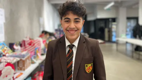 Youssef stands in front of a table full of selection boxes and boxes of sweets. He has short, dark curly hair. He wears a brown school blazer and a brown and orange striped tie
