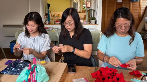 Three women sit in a row crocheting. They are making red Remembrance poppies and coasters. They are each holding crochet hooks and looking down at their work. 
