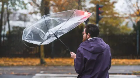 A man holds an upside down umbrella at the side of a road. He is wearing glasses and a purple raincoat.
