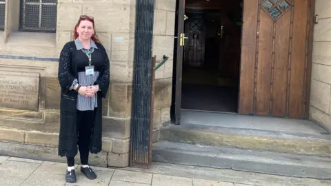 Sarah Porter. She is dressed in black and is standing outside the Hull and East Yorkshire Centre for the Deaf building. 