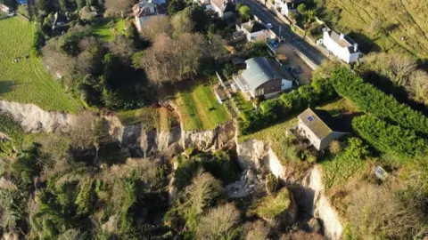 A drone image of a landslide on the Isle of Wight. The birdseye view shows a section of cliff, covered in grass has fallen down, with trees falling down the edge as well as what looks like a red shed. There is a house right on the edge of the cliff and the whole area is lined by trees.