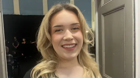 The head and shoulders of a young lady with long blonde hair smiling and looking at the camera. She is wearing pink lipstick and yellow eyeliner and is standing in front of a white door and some people dressed in black in a rehearsal space