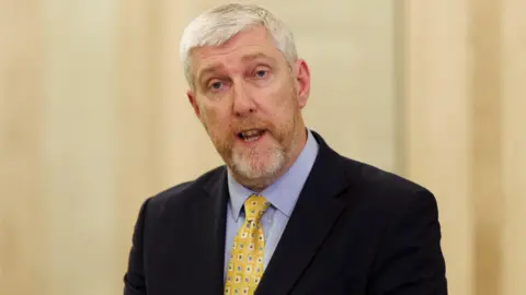 PA Media John speaking. He is wearing a blue shirt, yellow and blue patterned tie and a black suit jacket. He has grey hair.