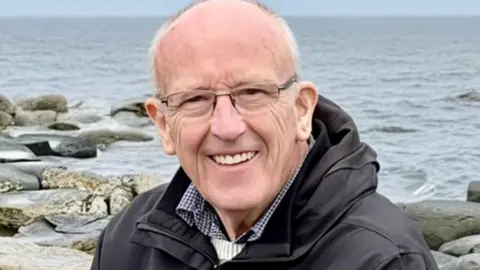 Edwin Williams Head and shoulders of Edwin Williams who sits on a rocky beach with a grey sea in the background. He is smiling, has very short white hair and glasses and is wearing a dark coat