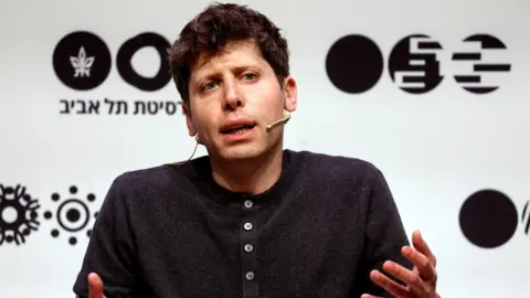 OpenAI leader Sam Altman sitting on a stage, speaking and gesturing with his hands while wearing a dark grey henley sweater.