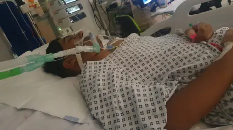 Ayyab Ayyub is ventilated and is lying down in a hospital bed wearing a hospital gown in ICU. He is surrounded by medical equipment