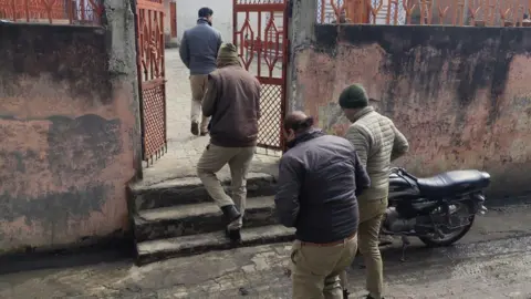 Four uniformed policemen enter the premises of a house in Umri village. Outside the gate of the house, a black motorbike can be seen parked