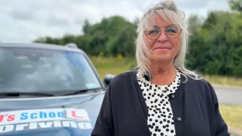 Sue Papworth looking directly at the camera dressed in a white and black spotty dress and black cardigan, with a driving school car behind her
