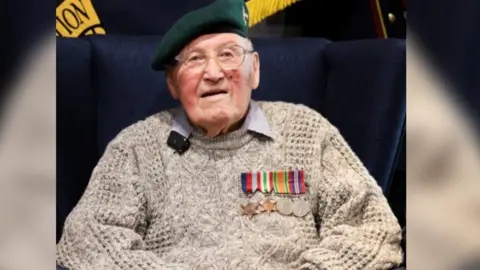 An elderly man man wearing a green beret, glasses, a knitted jumper and wearing a ribbon of medals sits in a chair.