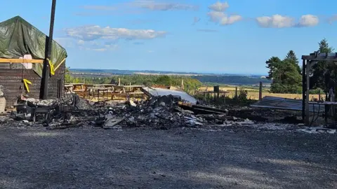 Remnants of the burnt wooden building are piled up. The structure was completely destroyed and another wooden building next to it is damaged with a tarpaulin over the roof.