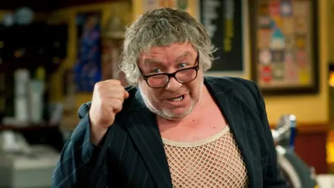 Gregor Fisher as Rab C Nesbitt. He is wearing a curly wig, black pinstripe blazer, glasses and string vest.