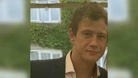 West Yorkshire Police A man with short brown hair smiles at the camera. He is wearing a suit and standing in front of a building covered in green leaves.