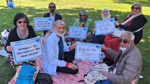 Supporting Humanity A group of older people sat on a picnic blanket. Some hold signs and they wave at the camera. It looks like a bright sunny day.