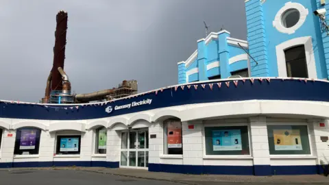 Exterior of Guernsey Electricity building with blue-and-white facade, arched windows displaying household-themed posters, and industrial chimney in background under overcast sky