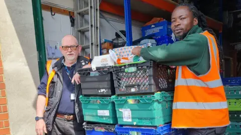 Luton Foodbank Two people standing by a large amount of boxes with food items in them. The boxes are black, green and blue. One man to the left is hearing a blue top, jacket, high-vis and trousers, the other man to the right is wearing a green top, dark trousers and a high-vis jacket. They are both looking at the camera.