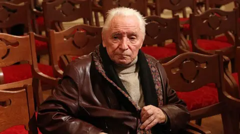 Grigorovich sits in a red seat at the theatre in a brown leather jacket